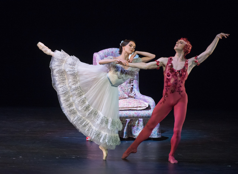 A ballerina in an ankle length white ruffled dress extends behind her while she holds the hand of her partner in a rose unitard